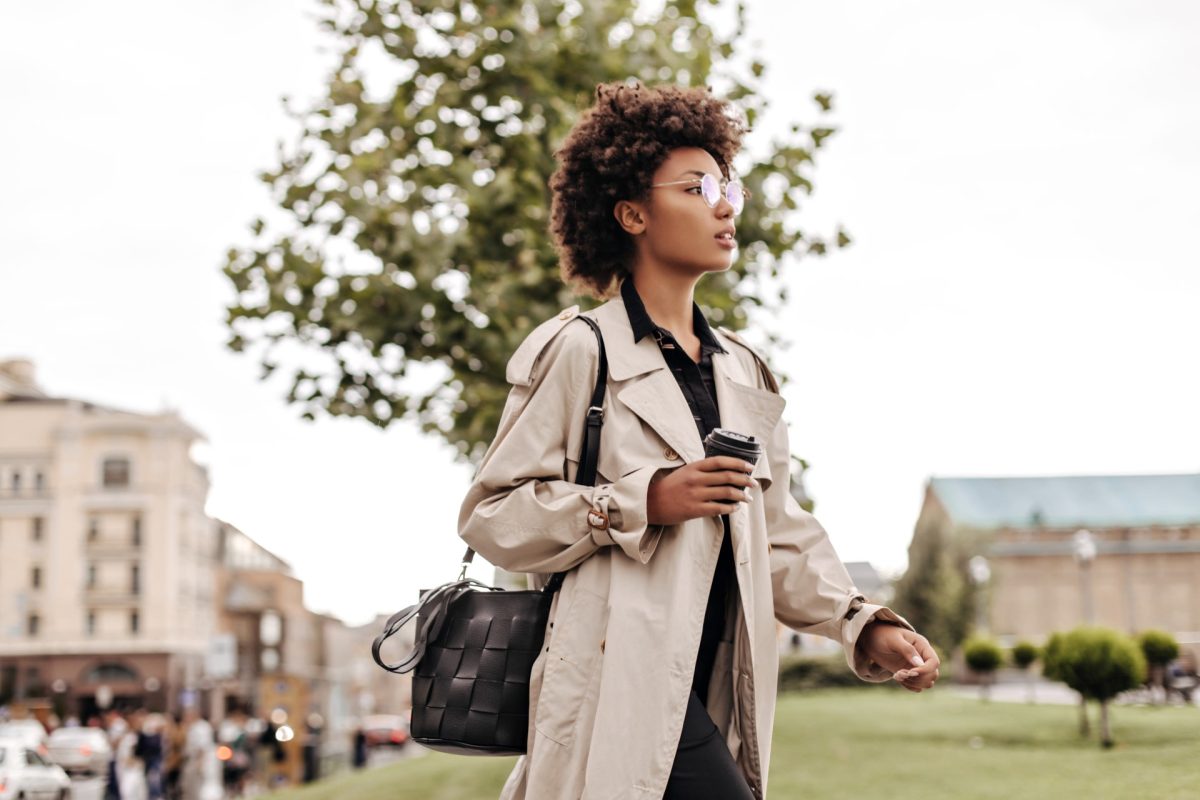 Attractive young brunette curly dark-skinned woman in black dress and beige trench coat walks outsi